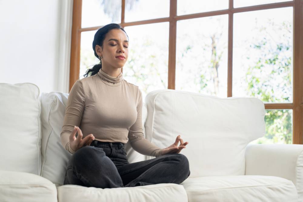A young woman practicing mindfulness meditation on a white couch, benefiting from holistic approaches in a Partial Care Program in New Jersey.
